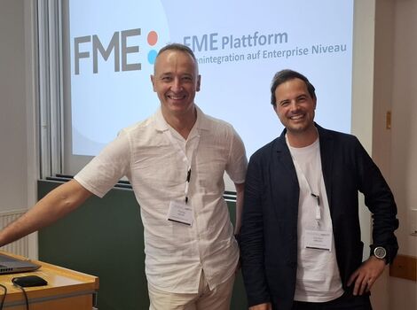 Zwei lächelnde Männer stehen in einem Schulungsraum vor einer Projektionswand mit dem FME-Logo und der Aufschrift „FME Plattform“. Sie tragen Namensschilder; einer in weißem Hemd und beiger Hose, der andere in dunkler Jacke und weißem T-Shirt.