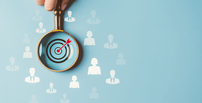 A hand holds a magnifying glass over a blue background, revealing a bullseye target with a red arrow hitting its center, symbolizing targeting a specific audience.