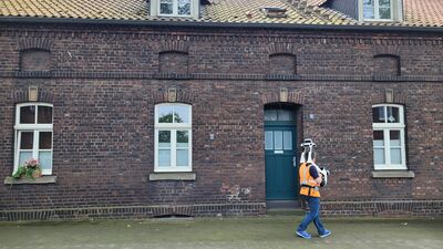 Eine Person mit orangefarbenem Rucksack geht an einer Backsteinfassade vorbei. Die Fassade hat eine blaue Tür und drei weiße Fenster, eines mit Blumenkästen. Die Szene wirkt ruhig; die Person trägt Jeans und Sneaker.